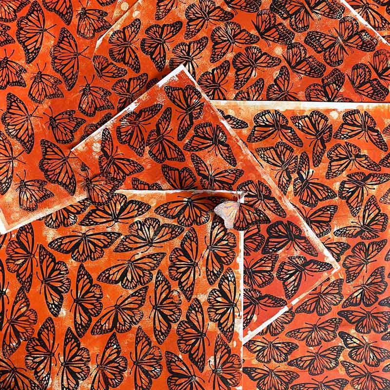 Overlapping sheets of orange paper are covered with black, detailed monarch butterfly patterns, creating a layered, vibrant, and textured design.