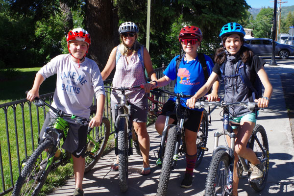 Four people, three youths and one adult, wearing helmets and casual clothes, stand outside on a sunny day with their bicycles, smiling at the camera near a fence and green trees.