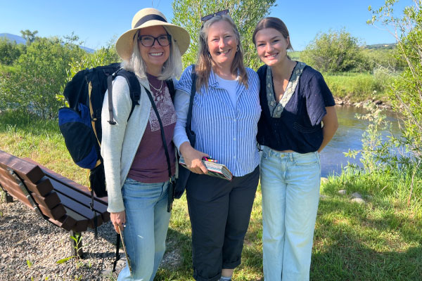 Three women stand outdoors smiling together near a stream. One woman wears a hat and backpack, another holds a notebook, and the third wears a dark top and light jeans. Trees, grass, and a wooden bench are in the background.
