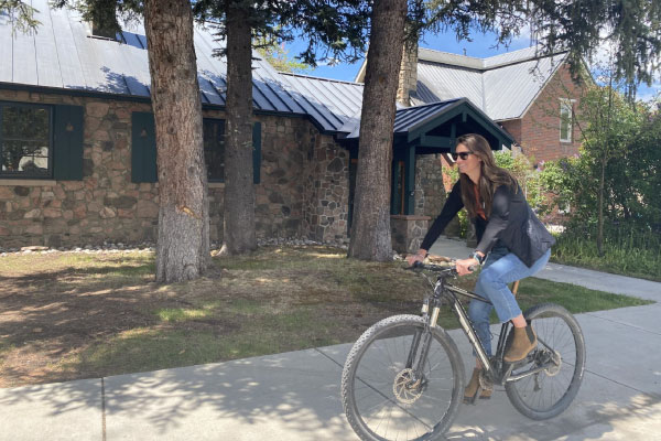A woman wearing sunglasses, a black jacket, jeans, and boots rides a bicycle on a sidewalk past a stone house with a metal roof, surrounded by trees on a sunny day.