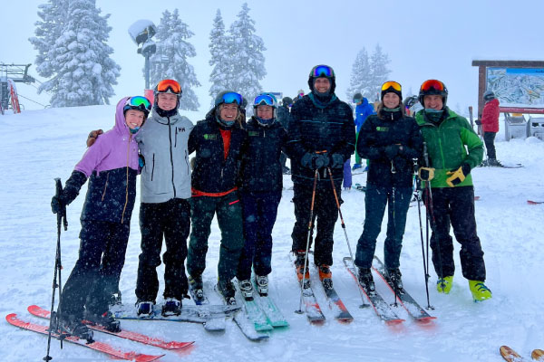 Seven people in colorful ski gear stand in a row on a snowy mountain slope, smiling at the camera with their skis on. Snow-covered trees and a ski map sign are visible in the background.