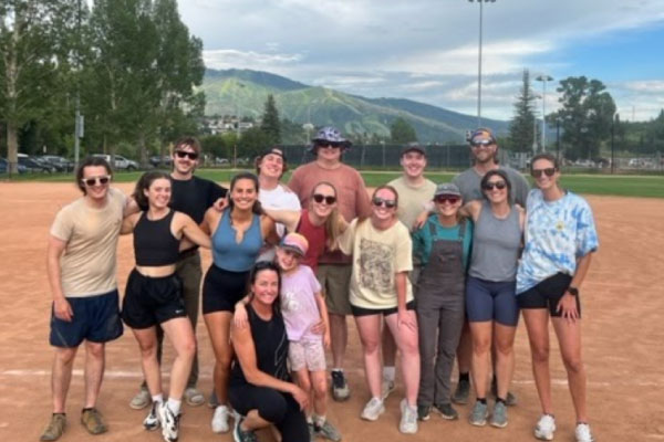 A group of fifteen people, including two children, pose and smile together on a baseball field with trees and green hills in the background on a cloudy day.