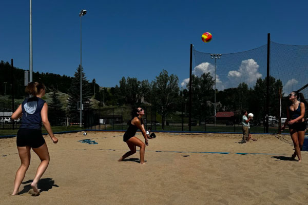Three women play beach volleyball on a sandy court under a clear blue sky, with one preparing to hit the ball. Trees, fencing, and two people standing in the background are visible.