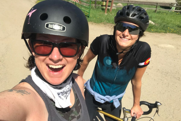 Two women wearing helmets and sunglasses smile at the camera while outdoors. One is holding a bike, and both are dressed in casual athletic gear on a sunny day.