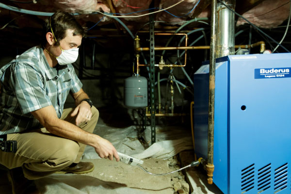 Western-Resilience-Center-energy-assessment A man wearing a mask and plaid shirt uses a handheld device to inspect a blue boiler in a dimly lit basement with exposed insulation and pipes.