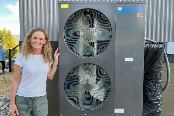 A smiling woman with curly hair stands next to a large outdoor heat pump unit labeled "ARCTIC" with two visible fans, in a sunny, outdoor setting.