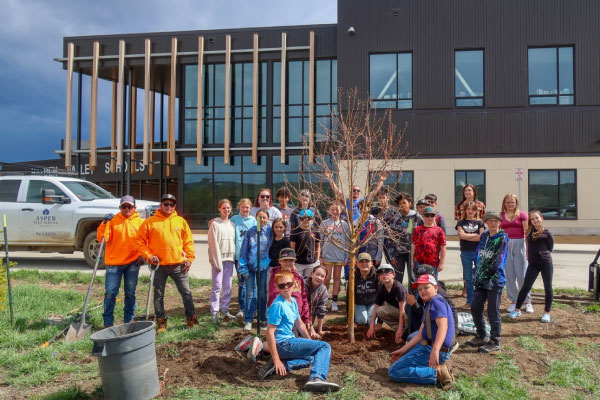 A group of children and adults gather around a newly planted tree in front of a modern building, smiling for the photo. Gardening tools and a truck are visible nearby on a sunny day.