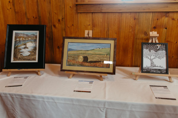 Three framed artworks are displayed on small easels on a white-covered table against a wooden wall. The pieces feature a river scene, a rural landscape with a hay bale, and a stylized tree with a crescent moon.