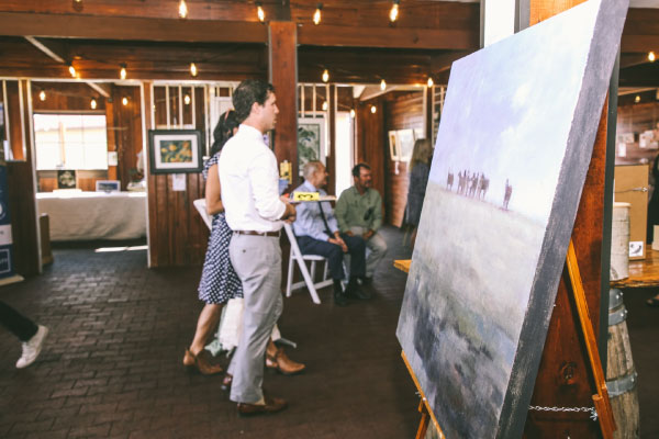 People stand and observe a large painting on an easel at an art gallery or exhibition, with other artworks displayed on the wooden walls and several people seated in the background.