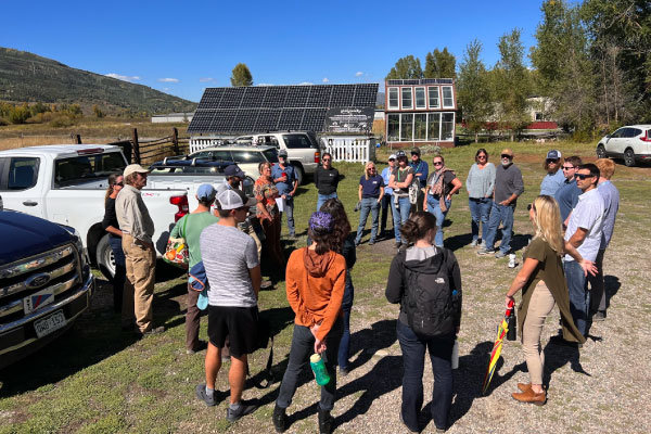 A group of people stands in a circle outdoors near parked vehicles, with solar panels and a small building in the background on a sunny day. Trees and hills are visible under a clear blue sky.