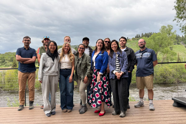 A group of eleven people stands smiling on a wooden deck outdoors, with a river, green trees, and hills in the background under a cloudy sky.
