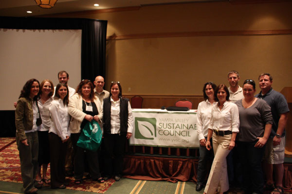 A group of thirteen people stands in a conference room beside a table with a banner reading "Yampa Valley Sustainable Council." They are smiling at the camera, and the room is softly lit.