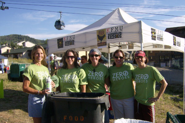 Five women wearing matching green "Zero Heroes" shirts stand and smile in front of a tent with "KFMU Radio" signs at an outdoor event, with mountains and gondolas visible in the background.