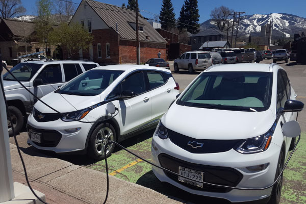 Two white Chevrolet electric cars are parked and charging at a station in a small town. Snow-capped mountains and houses are visible in the background under a clear sky.