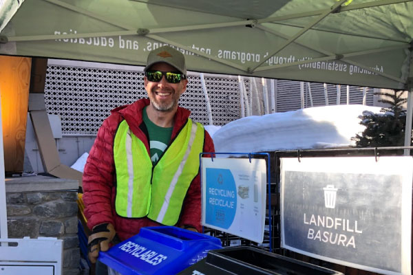 A smiling person wearing sunglasses, a cap, and a reflective vest stands outdoors under a tent, holding a blue recycling bin next to a landfill bin labeled "Landfill Basura.