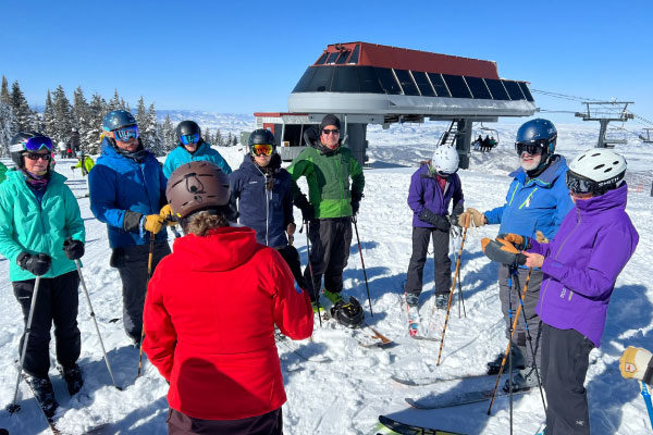 A group of skiers in winter gear stand in a circle on a snowy slope near a ski lift, talking and holding ski poles, with clear blue sky and trees in the background.