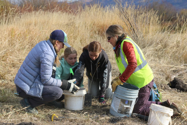 Four people kneel on dry grass in an open field, examining buckets together. One wears a reflective vest, and the others wear jackets and hats. Tall grass and distant trees are visible in the background.