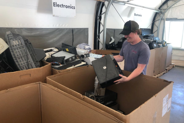 A person smiles while placing old electronic devices into a large cardboard box labeled for electronics recycling inside a collection facility.
