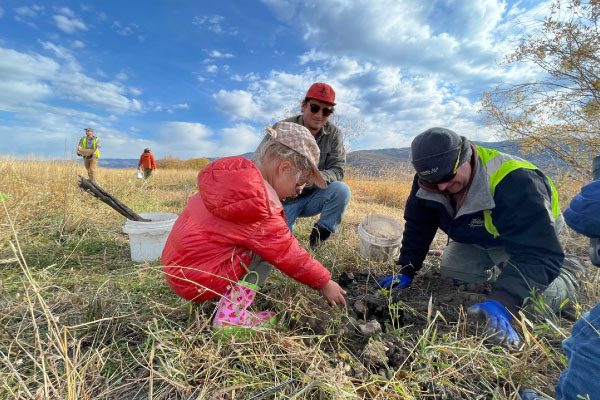 A young girl in a red jacket and two adults dig in the soil on a grassy field under a blue sky. Other people work in the background, and buckets and tools are scattered nearby.