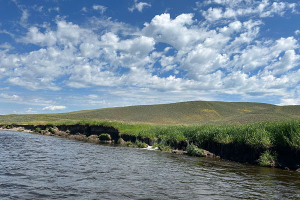 A calm river flows alongside grassy banks under a blue sky with scattered white clouds, with rolling green hills in the background.