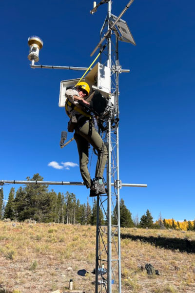 A person wearing safety gear climbs a tall metal weather tower in a grassy field, adjusting equipment under a clear blue sky with trees in the background.