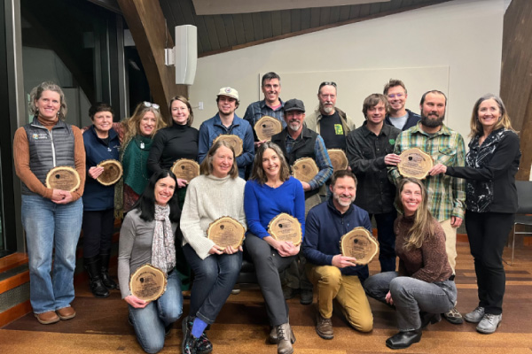 A group of 17 people pose indoors, each holding a wooden plaque shaped like a tree slice, smiling at the camera, suggesting an awards or recognition event.