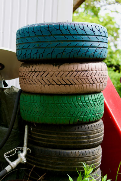 A stack of four tires painted blue, pink, green, and black, standing outdoors near a white building, with some plants and objects visible around them.