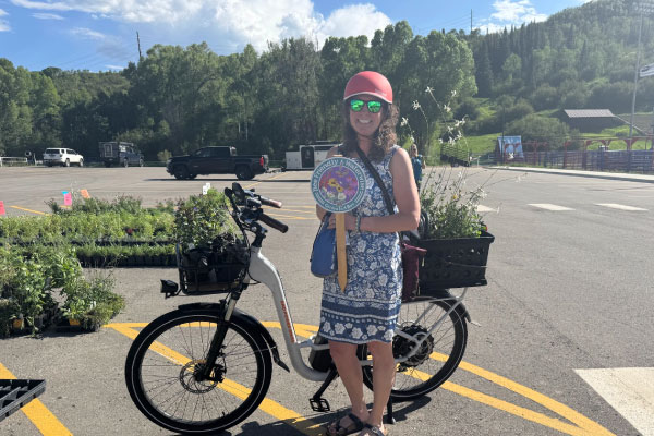 A woman wearing a red helmet stands next to an electric bike with baskets full of plants. She is smiling and holding a colorful circular sign. Trees and a parking area are visible in the background.