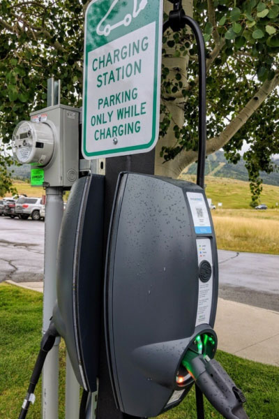 An electric vehicle charging station with a plugged-in charger sits beneath a sign that reads, “Charging station, parking only while charging.” Trees, grass, and parked cars are visible in the background.