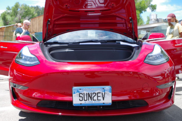 A red electric car with its front trunk open displays a Colorado license plate reading "SUN2EV." Several people stand nearby, and trees and a building are visible in the background.