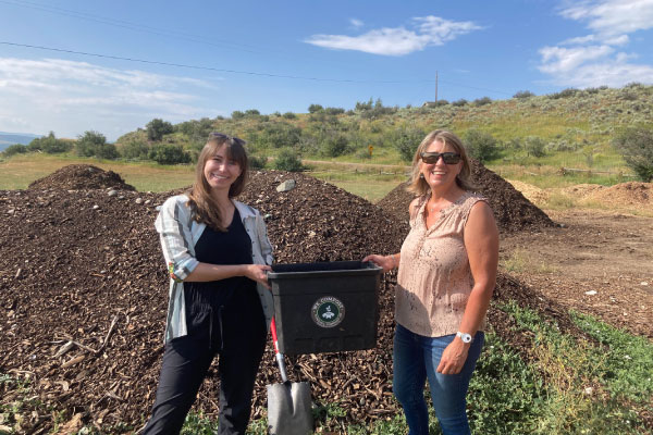 Two women stand outdoors in front of mulch piles, smiling and holding a black compost bin together. One holds a shovel. It is a sunny day with blue sky and greenery in the background.