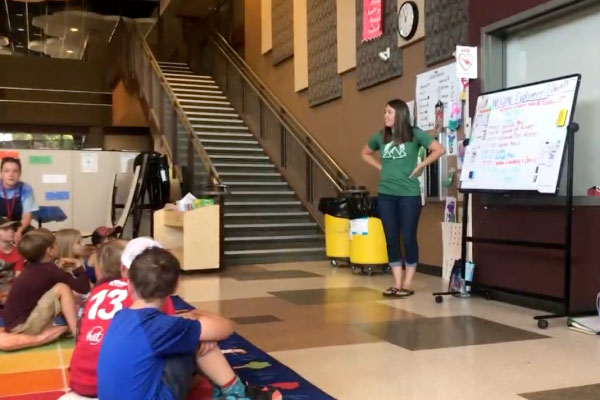 A woman stands near a whiteboard, speaking to a group of children seated on a colorful rug in a school hallway. There is a staircase, bulletin boards, and a large clock in the background.