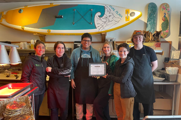 Six smiling restaurant staff members stand together indoors, one holding a framed certificate. Behind them is a large yellow paddleboard mounted on the wall, along with surf-themed décor.