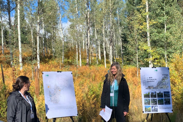 Two women stand outdoors in front of easels displaying maps and images, surrounded by tall trees and autumn foliage, giving a presentation in a natural setting under a sunny sky.