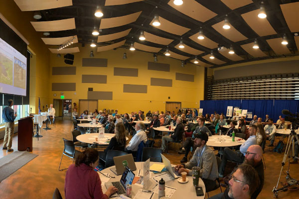 A large group of people sit at round tables in a spacious conference room, listening to a speaker at a podium. Presentation slides are projected on a screen at the front of the room.