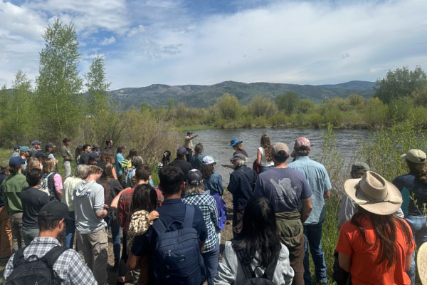 A large group of people stands by a riverbank listening to a speaker, surrounded by green shrubs and trees, with mountains and a blue sky with clouds in the background.