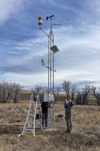 Two people work on a tall weather station tower in a grassy field; one stands on a ladder adjusting equipment, while the other stands nearby. Leafless trees and a cloudy sky are in the background.
