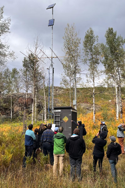 A group of people stand in a grassy field, observing a weather station with antennas and a solar panel, surrounded by tall trees and autumn foliage under a cloudy sky.