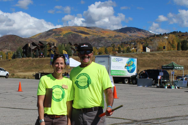 Two people wearing bright yellow "Zero Hero" shirts stand smiling in an outdoor lot, with mountains and trees displaying autumn colors in the background under a blue sky with clouds.