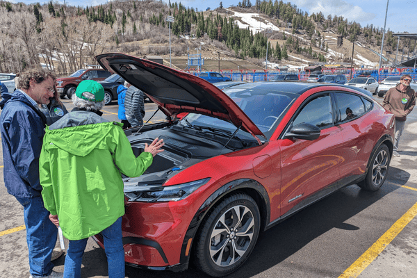 Image of a man in a green rain coat talking to a man in a blue raincoat in front of a red EV with the hood up.