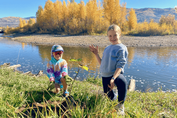 Image of two kids showing off a tree sapling they just planted, river in background.