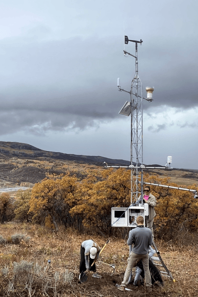 Image of a YBASIN station, a tall set of poles and measuring devices, in a field, with two people working in front of it.