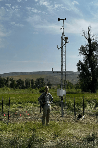Image of a YBASIN station, a tall set of poles and measuring devices, in a field, with a person posing in front of it.