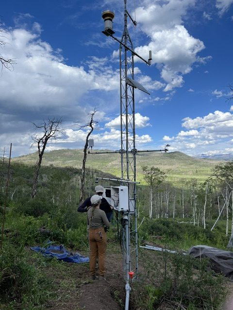 Photo of a soil moisture monitoring station with people standing working on it and mountains and forest in the background.